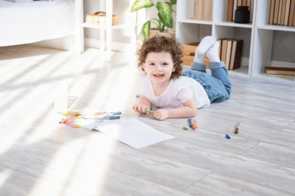 Child lying on a light floor, drawing with colorful markers on paper in a bright, tidy living space.