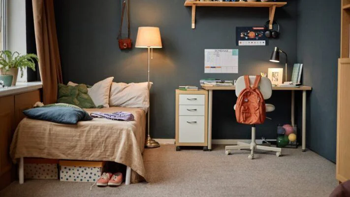 A cozy dorm-style bedroom with a single bed on the left, a desk and chair against a dark wall, and a bright orange backpack hanging on the chair.