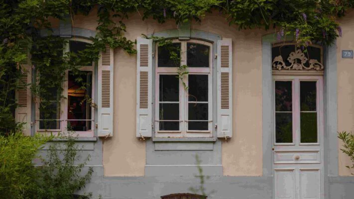 Beige house facade with gray trim, ivy over the roof, three windows with white shutters, and a white door on the right (no. 79).