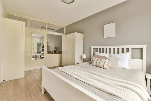 White wooden bed with beige bedding and decorative pillows in a neutral gray-walled bedroom.